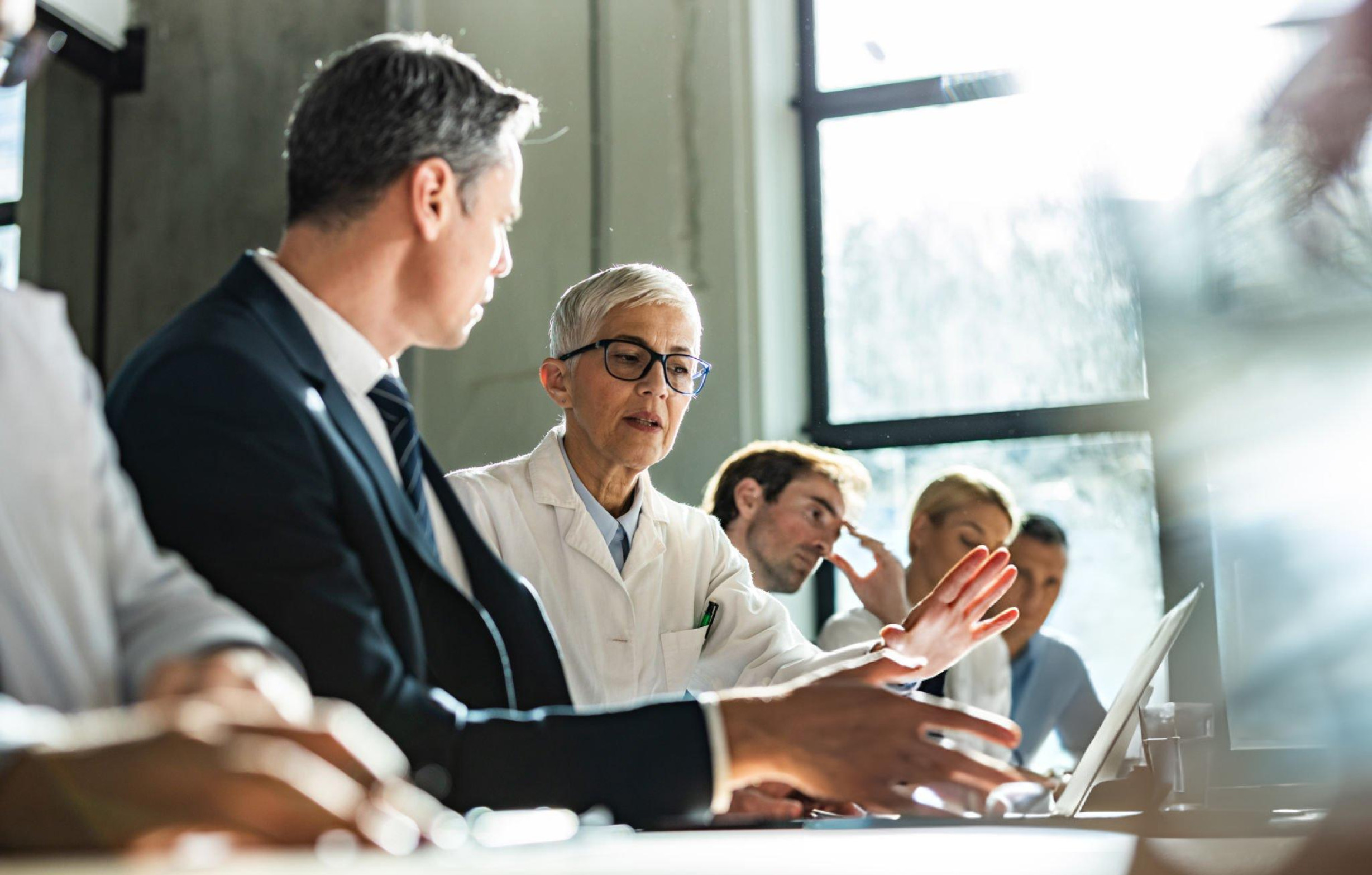 A imagem mostra um grupo de profissionais em uma reunião de negócios, com foco principal em duas pessoas no centro da imagem. No primeiro plano e à esquerda, um homem de terno escuro está sentado, olhando atentamente para uma mulher à sua direita. Esta mulher, de cabelo curto e grisalho, usa óculos e um jaleco branco com detalhes verdes, indicando que ela pode ser uma profissional da área da saúde, como uma médica ou pesquisadora. Ela gesticula com a mão esquerda aberta enquanto parece explicar ou apresentar algo em um laptop que está à frente dela. Outras pessoas estão sentadas à mesa ao fundo, algumas visivelmente prestando atenção à discussão, enquanto o ambiente é iluminado pela luz natural que entra por uma grande janela ao fundo, criando um efeito de contraluz em parte da cena, representando uma conversa sobre marketing médico