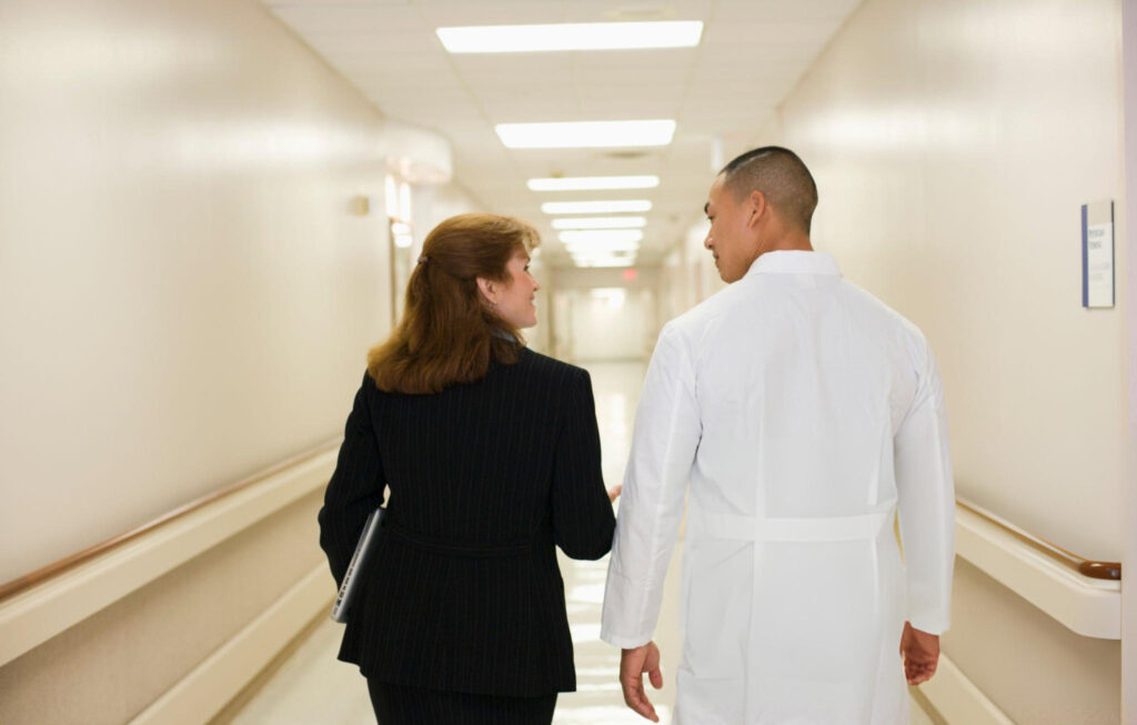Duas pessoas caminhando lado a lado em um corredor de hospital ou instituição de saúde. À esquerda, há uma mulher vestida com um terno escuro e segurando um notebook; à direita, um homem usando um jaleco branco, possivelmente um médico. Ambos parecem conversar enquanto caminham, transmitindo uma sensação de profissionalismo, colaboração no ambiente hospitalar e bate-papo sobre marketing médico.