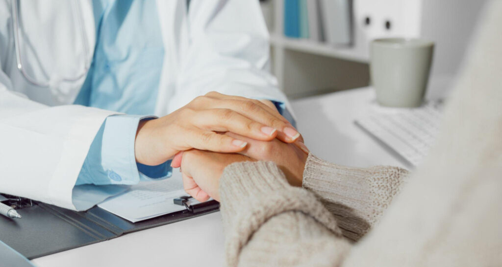 A imagem mostra um gesto de empatia e cuidado entre profissional de saúde e paciente. Um médico (ou médica) segura com as duas mãos as mãos de uma pessoa, que veste um suéter bege, em sinal de apoio. O profissional usa jaleco branco e camisa azul-clara, sentado à mesa com papéis e caneta, sugerindo um ambiente de consultório. Ao fundo, há itens de escritório, como teclado e caneca, reforçando o contexto clínico. Mensagem visual: representa confiança, acolhimento e vínculo humano na relação médico-paciente, o toque transmite segurança e escuta ativa durante a Jornada do Paciente.