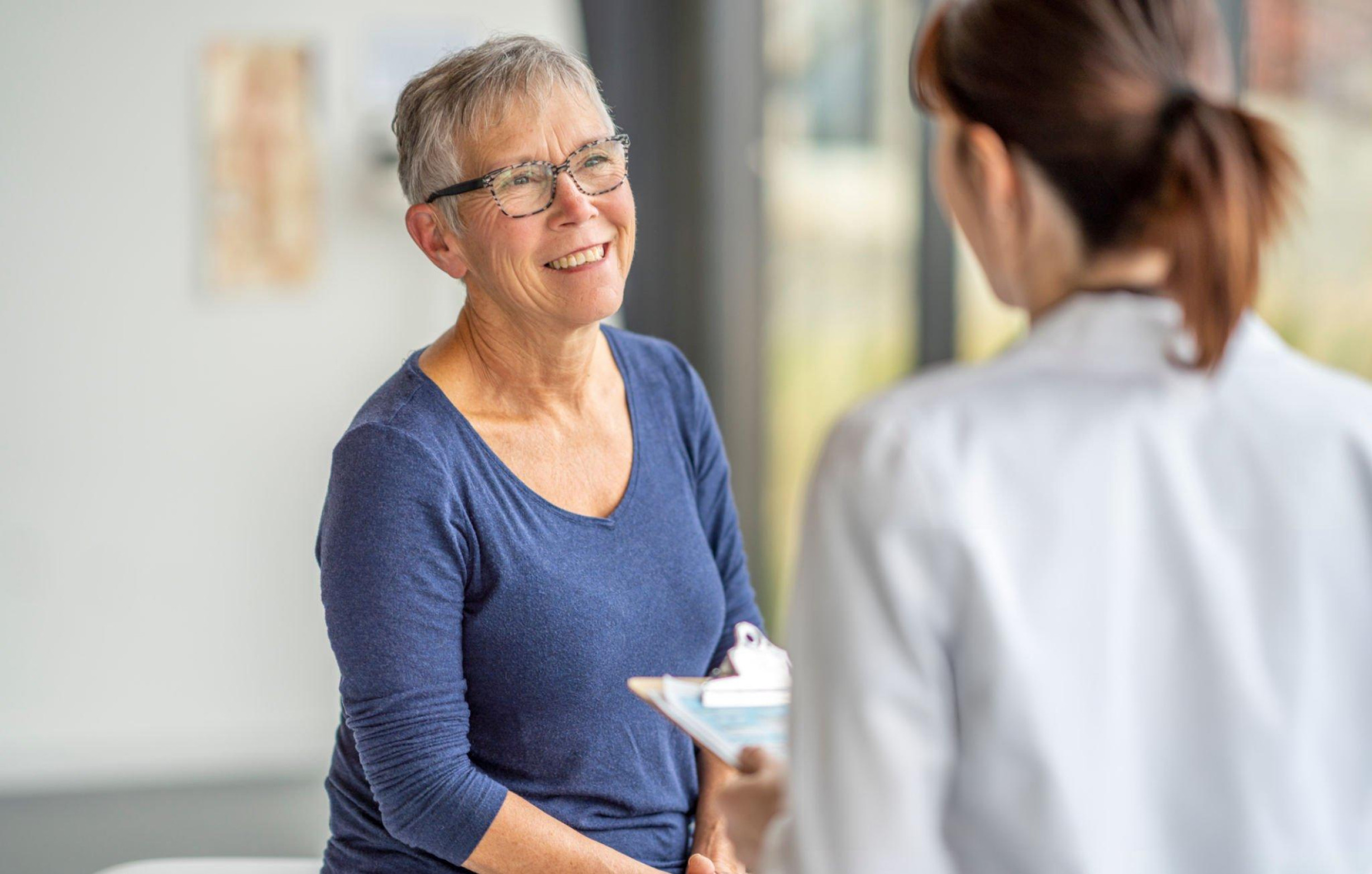 A foto mostra uma paciente idosa sorridente conversando com uma profissional de saúde (provavelmente médica ou enfermeira). A paciente, de cabelos curtos e grisalhos, usa óculos e uma blusa azul. Ela está sentada, demonstrando tranquilidade e confiança durante o atendimento. A profissional aparece de costas, com jaleco branco e prancheta nas mãos, indicando que está ouvindo atentamente ou anotando informações. O ambiente é claro e acolhedor, com fundo neutro e janelas que permitem entrada de luz natural. Mensagem visual: transmite empatia, acolhimento e uma boa relação médico-paciente, reforçando a importância da comunicação durante a Jornada do Paciente.
